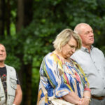 Trzy osoby stoją w lesie, patrząc w dół. Kobieta ma kolorową bluzkę, mężczyźni szare ubrania. Tło jest zielone i naturalne.