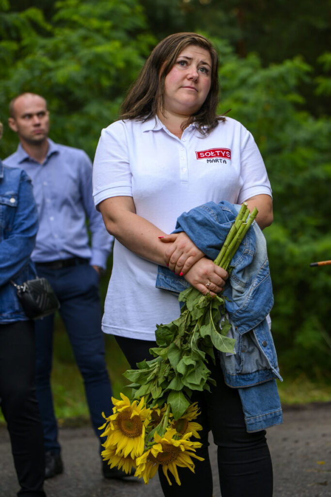 Kobieta w białej koszulce trzyma słoneczniki i kurtkę jeansową. W tle zielone drzewa i dwie osoby. Jest na zewnątrz na chodniku.