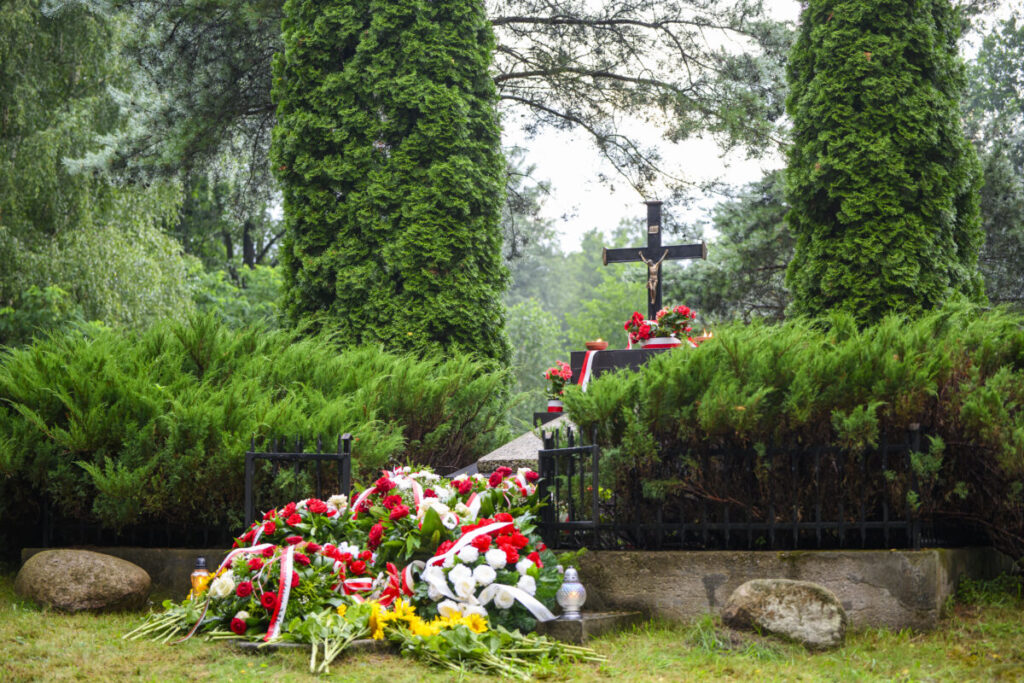 Na zdjęciu widoczny jest zadbany grób ozdobiony kwiatami w czerwieni i bieli. Tło stanowi zieleń drzew. Na pomniku znajduje się krzyż.