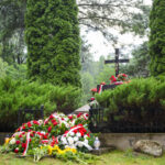 Na zdjęciu widoczny jest zadbany grób ozdobiony kwiatami w czerwieni i bieli. Tło stanowi zieleń drzew. Na pomniku znajduje się krzyż.