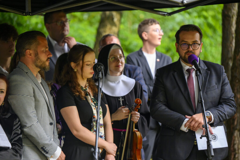 Grupa osób stoi pod namiotem; mężczyzna w garniturze przemawia. Tło jest zielone i zalesione. Obecna jest zakonnica z instrumentem.