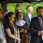 Grupa osób stoi pod namiotem; mężczyzna w garniturze przemawia. Tło jest zielone i zalesione. Obecna jest zakonnica z instrumentem.