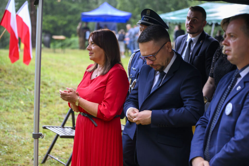 Grupa ludzi na zewnątrz, kobieta w czerwonej sukience, mężczyźni w garniturach. W tle zielona trawa i polskie flagi.