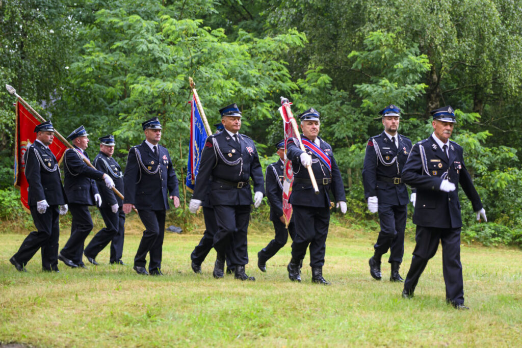 Mężczyźni w czarnych mundurach maszerują z flagami po trawniku, otoczeni zielenią drzew.