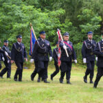 Mężczyźni w czarnych mundurach maszerują z flagami po trawniku, otoczeni zielenią drzew.