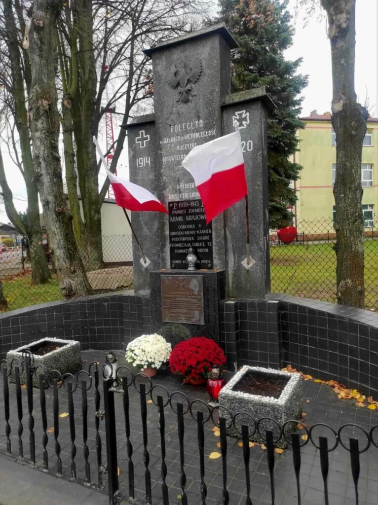 Pomnik z szarym krzyżem i polskimi flagami. Obok biało-czerwone chryzantemy. Tło to drzewa i ogrodzenie.