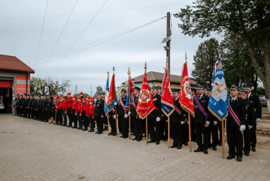 Grupa strażaków stoi w szeregu z czerwonymi i niebieskimi sztandarami. Ubrani są w czarne mundury i czerwone kurtki. Tło stanowi budynek z czerwonym dachem i drzewa.