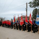 JTba5aGF Grupa strażaków stoi w szeregu z czerwonymi i niebieskimi sztandarami. Ubrani są w czarne mundury i czerwone kurtki. Tło stanowi budynek z czerwonym dachem i drzewa.