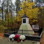 Pomnik otoczony czarnym ogrodzeniem z kolorowymi chryzantemami i zniczami. Tło leśne z żółtymi i brązowymi liśćmi. Jesienna atmosfera.