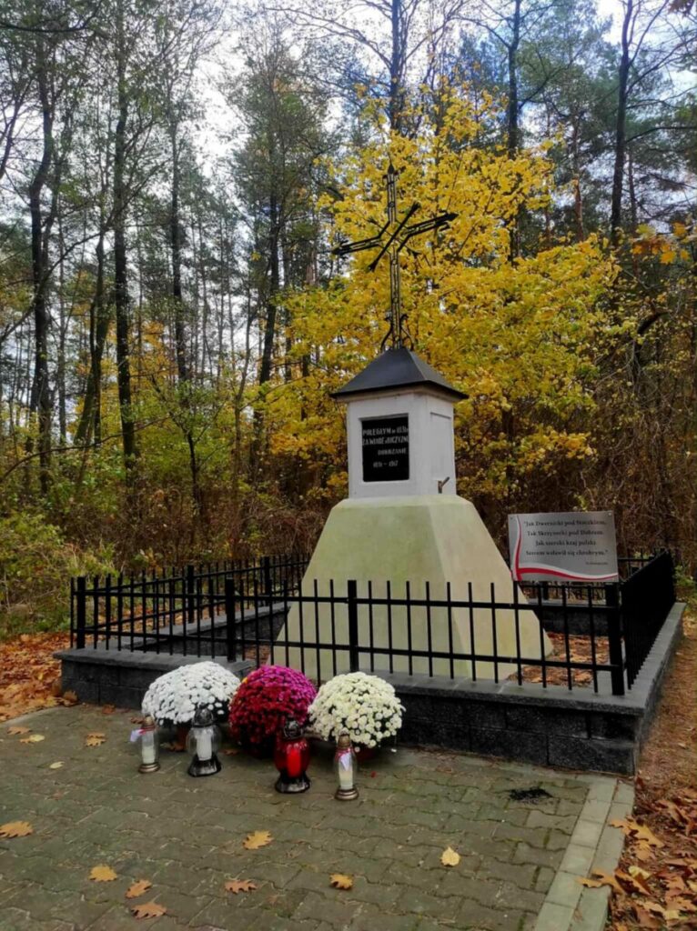 Pomnik otoczony czarnym ogrodzeniem z kolorowymi chryzantemami i zniczami. Tło leśne z żółtymi i brązowymi liśćmi. Jesienna atmosfera.