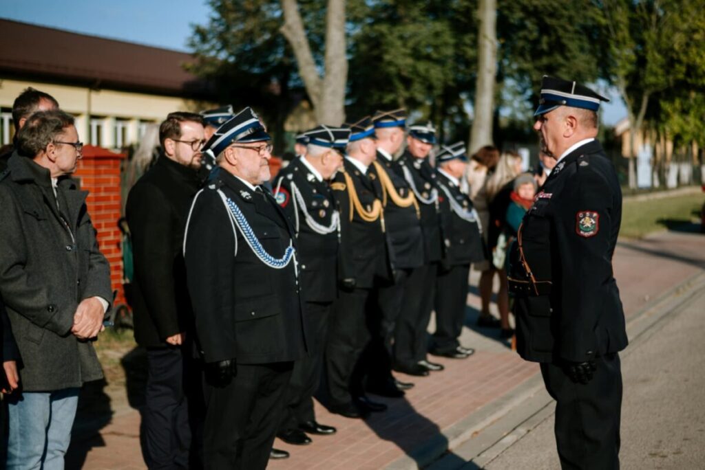Grupa osób w mundurach strażackich stoi na chodniku. Obok nich kilka osób w cywilnych ubraniach. W tle widać drzewa i budynek.