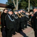 N58UqMbA Grupa osób w mundurach strażackich stoi na chodniku. Obok nich kilka osób w cywilnych ubraniach. W tle widać drzewa i budynek.