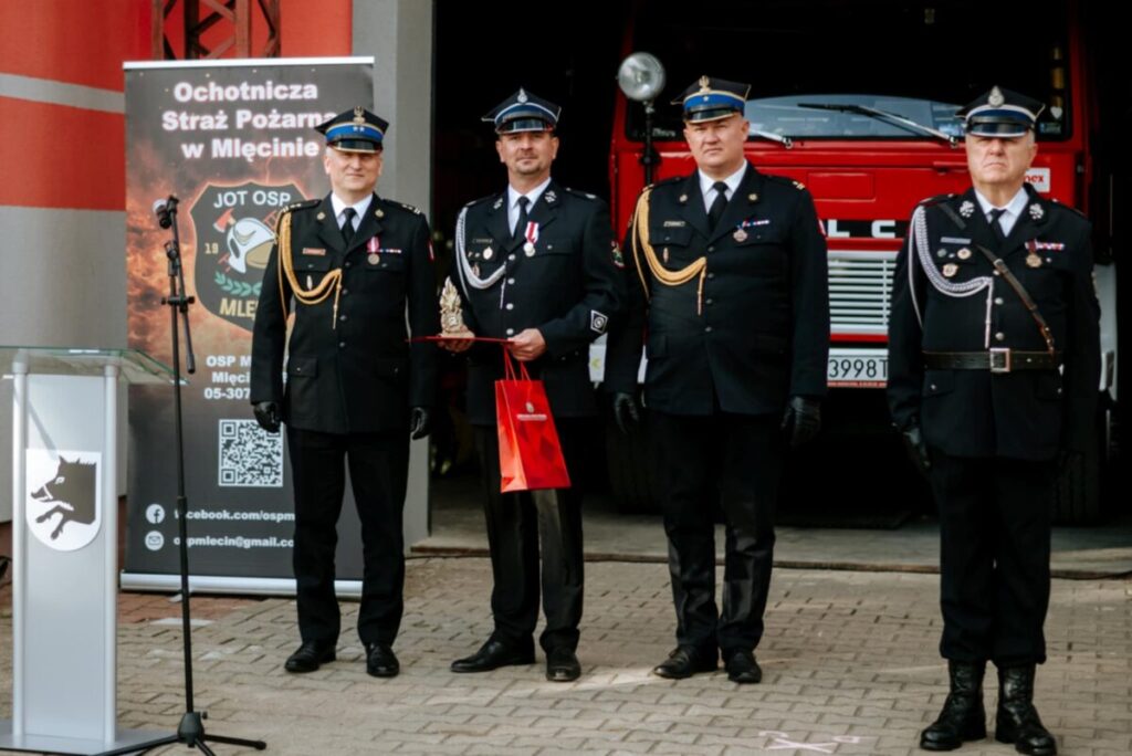 Czterech strażaków w ciemnych mundurach stoi przed czerwonym wozem strażackim. Dwóch z nich trzyma nagrody. W tle jest transparent OSP.