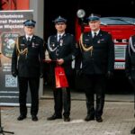 NJDAjZan Czterech strażaków w ciemnych mundurach stoi przed czerwonym wozem strażackim. Dwóch z nich trzyma nagrody. W tle jest transparent OSP.