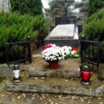 Grób z czarnym krzyżem otoczony iglakami. Na schodach biało-czerwone kwiaty i znicze. W tle drzewa.