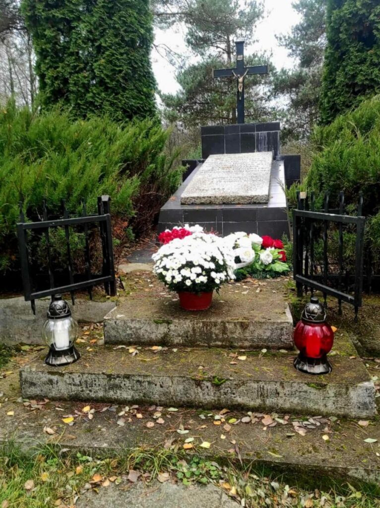 Nagrobek z krzyżem na cmentarzu, wokół kwiaty i znicze w tle zielone drzewa.