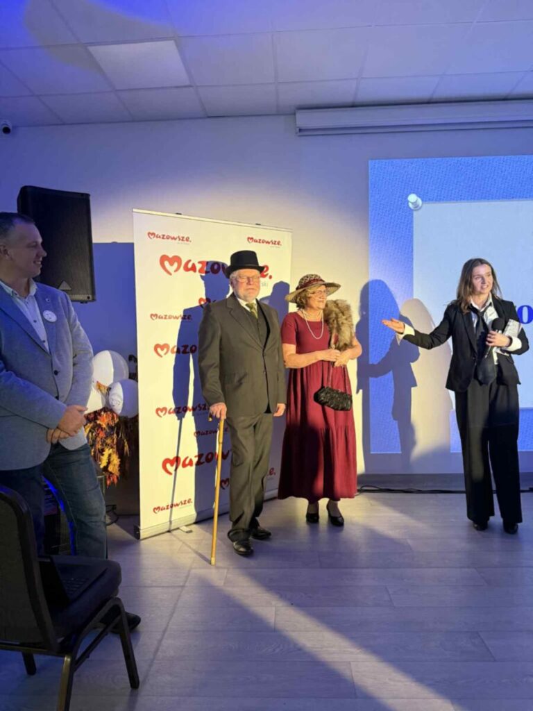 Na zdjęciu widać cztery osoby na jasnym tle. Trzy z nich stoją na scenie, dwie trzymając mikrofon. Tło zdobi napis Mazowsze.