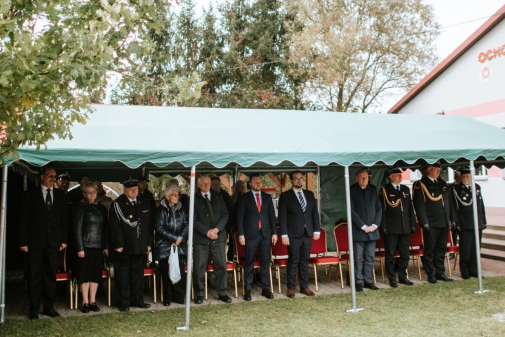 Grupa ludzi w eleganckich strojach stoi pod zielonym namiotem na trawie. Tło zdobią drzewa i budynek OSP. Dominuje kolor zielony.