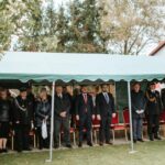 WxrONUo9 Grupa ludzi w eleganckich strojach stoi pod zielonym namiotem na trawie. Tło zdobią drzewa i budynek OSP. Dominuje kolor zielony.