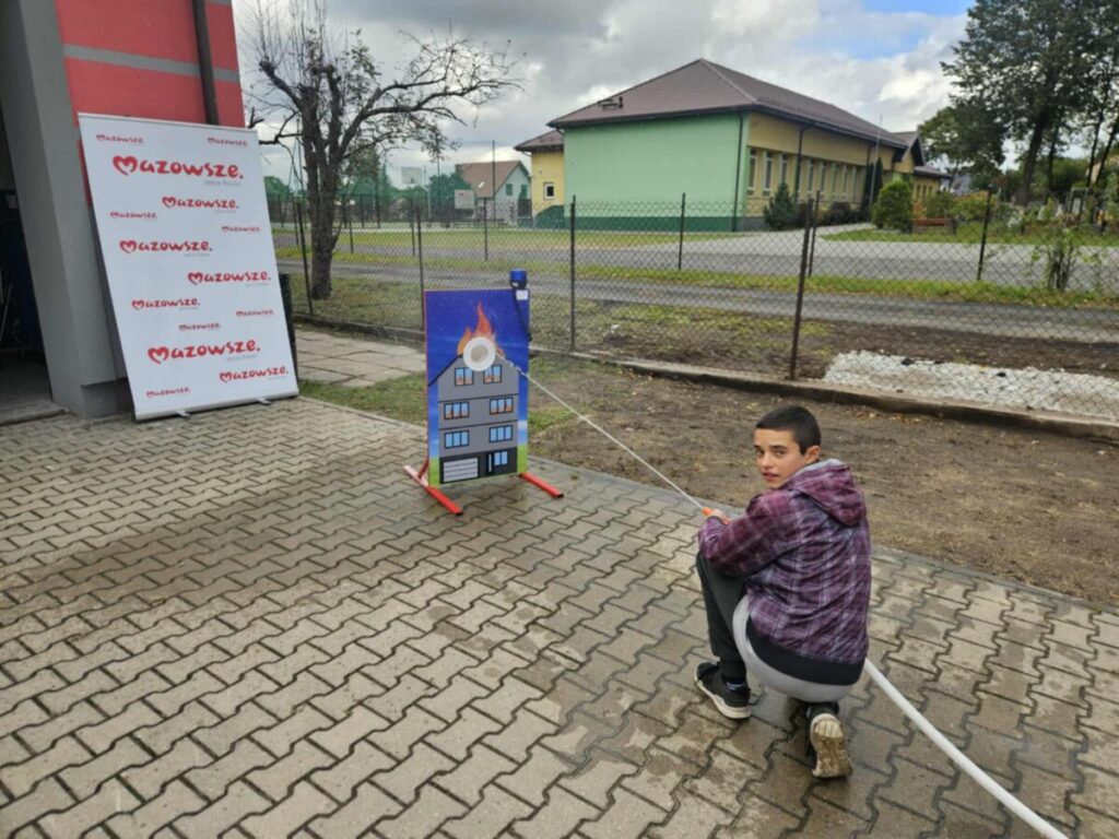 Młody chłopiec obsługuje wąż strażacki przy planszy z atrapą płonącego budynku. W tle zielony budynek i ogrodzenie. Pogoda pochmurna.