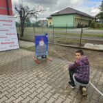 XNT6gdNN Młody chłopiec obsługuje wąż strażacki przy planszy z atrapą płonącego budynku. W tle zielony budynek i ogrodzenie. Pogoda pochmurna.