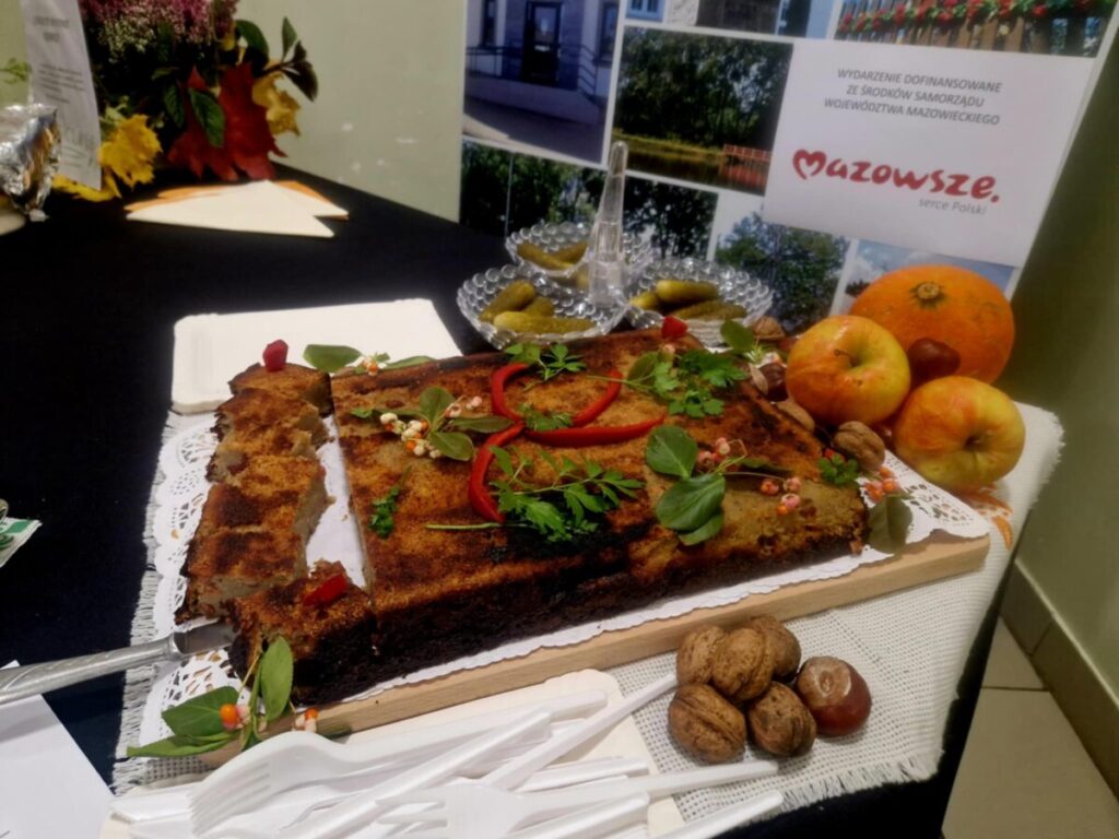 Na stole ciasto z ziołami i papryką, obok jabłka i orzechy. W tle ciemny obrus, ozdoby z liści i plakat Mazowsza.