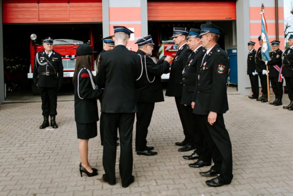 Grupa strażaków w czarnych mundurach stoi przed remizą z czerwonymi drzwiami. W tle widoczny jest czerwony wóz strażacki. Strażacy uczestniczą w ceremonii na placu z kostką brukową.