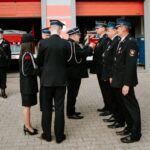 -dLaqwOK Grupa strażaków w czarnych mundurach stoi przed remizą z czerwonymi drzwiami. W tle widoczny jest czerwony wóz strażacki. Strażacy uczestniczą w ceremonii na placu z kostką brukową.