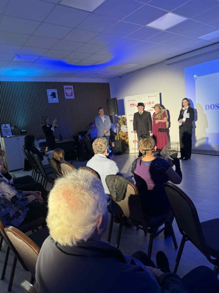 Grupa ludzi w sali konferencyjnej na tle niebieskiej ściany. Mężczyzna i kobieta w eleganckich strojach na scenie. Publiczność siedzi przodem do sceny, niektóre osoby robią zdjęcia.