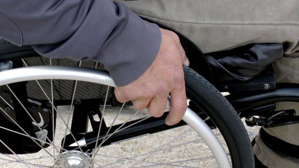 Osoba w szarej kurtce trzyma wózek inwalidzki. Tło jest rozmyte, dominuje szarość i czerń. Ręka skupiona na metalowym kole.