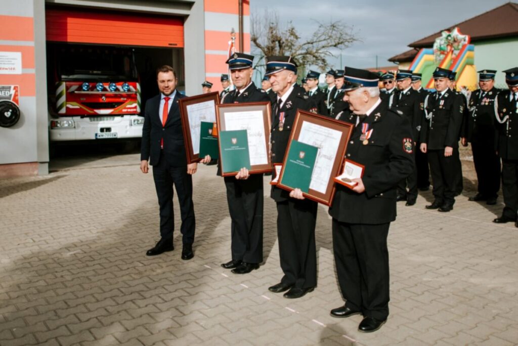 Czterej strażacy w mundurach z medalami i dyplomami stoją przed strażnicą. W tle wóz strażacki i grupa strażaków. Mężczyzna w garniturze po lewej stronie.