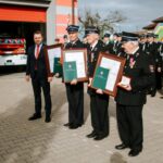 i4q3AxT8 Czterej strażacy w mundurach z medalami i dyplomami stoją przed strażnicą. W tle wóz strażacki i grupa strażaków. Mężczyzna w garniturze po lewej stronie.