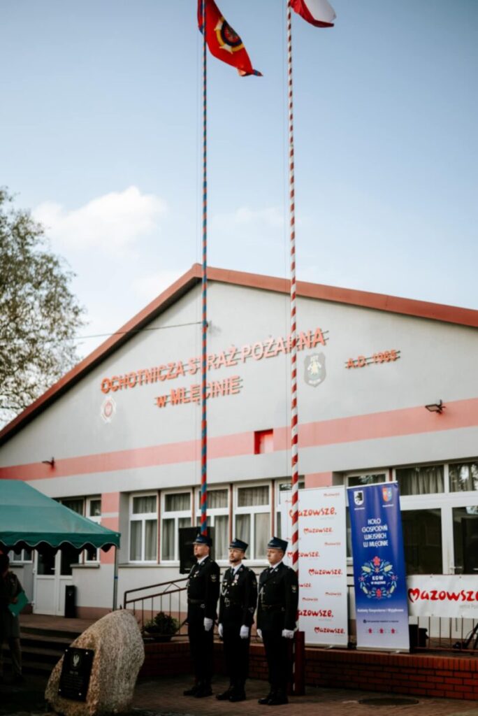 Czterech strażaków stoi przed budynkiem straży pożarnej z biało-czerwonymi flagami. Budynek jest szary z czerwonymi detalami. Tło stanowi niebieskie niebo z chmurami.