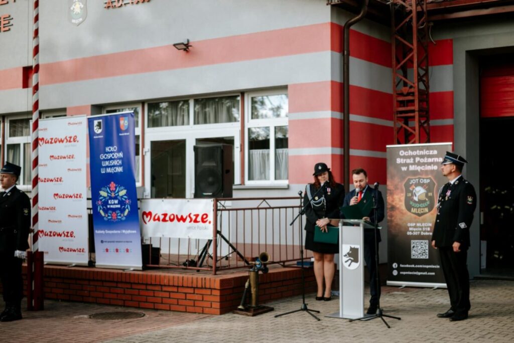 Grupa ludzi w mundurach strażackich stoi przed budynkiem z czerwono-szarymi pasami. Jedna osoba przemawia do mikrofonu. Obok znajdują się banery z napisem Mazowsze.