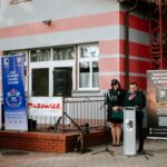 jF3nugvU Grupa ludzi w mundurach strażackich stoi przed budynkiem z czerwono-szarymi pasami. Jedna osoba przemawia do mikrofonu. Obok znajdują się banery z napisem Mazowsze.