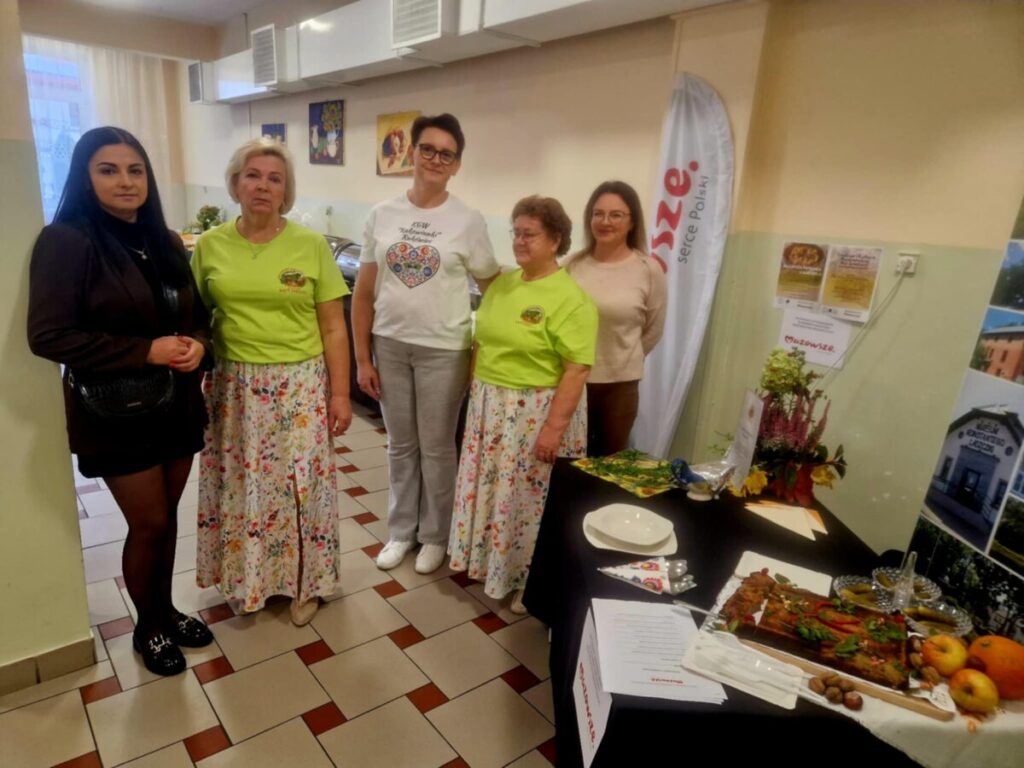 Na zdjęciu znajduje się pięć osób w jasnym pomieszczeniu. Kobiety mają na sobie kolorowe ubrania i stoją obok stołu. Na stole leży jedzenie i dekoracje, w tle widać plakaty i flagę.