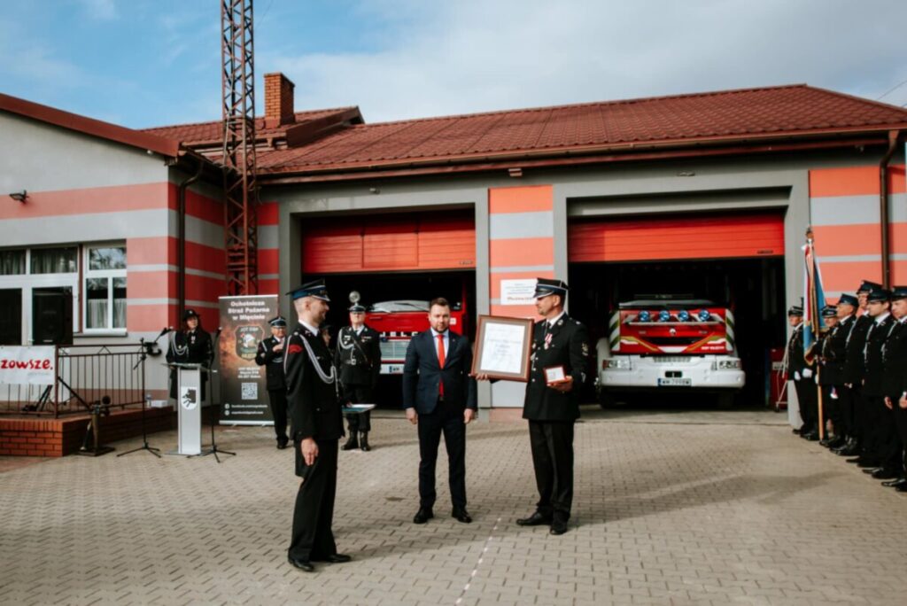 Na zdjęciu widać strażaków przed budynkiem OSP o szarej i czerwonej kolorystyce. W tle zaparkowany jest wóz strażacki. Dwóch strażaków wręcza certyfikat mężczyźnie w garniturze.