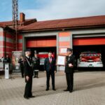 qS5_J3eT Na zdjęciu widać strażaków przed budynkiem OSP o szarej i czerwonej kolorystyce. W tle zaparkowany jest wóz strażacki. Dwóch strażaków wręcza certyfikat mężczyźnie w garniturze.
