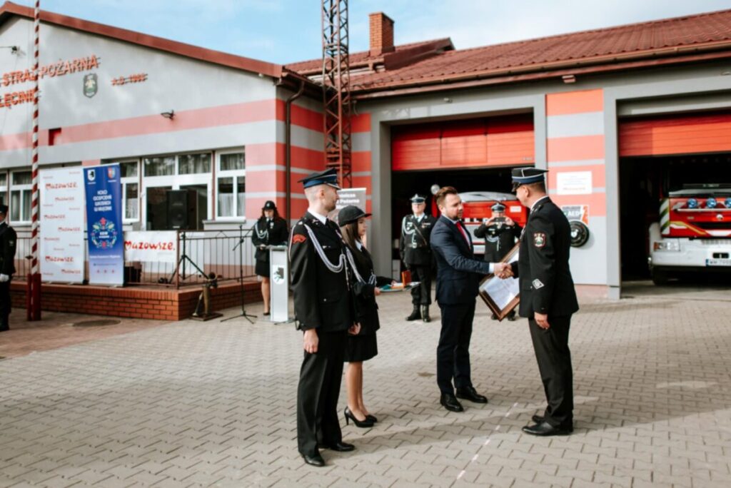 Grupa strażaków stoi przed remizą, odbierając nagrodę. Budynek jest szaro-czerwony, z otwartymi drzwiami garażu. W tle widoczne są pojazdy strażackie.