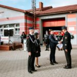 rJj5Y9Mj Grupa strażaków stoi przed remizą, odbierając nagrodę. Budynek jest szaro-czerwony, z otwartymi drzwiami garażu. W tle widoczne są pojazdy strażackie.