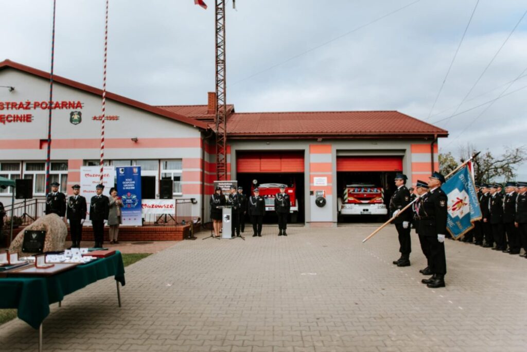 Na zdjęciu remiza strażacka z czerwonymi drzwiami i flagami. Strażacy w mundurach stoją przodem na placu. Tło z chmurami.