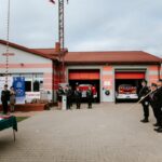 rr8vlijE Na zdjęciu remiza strażacka z czerwonymi drzwiami i flagami. Strażacy w mundurach stoją przodem na placu. Tło z chmurami.