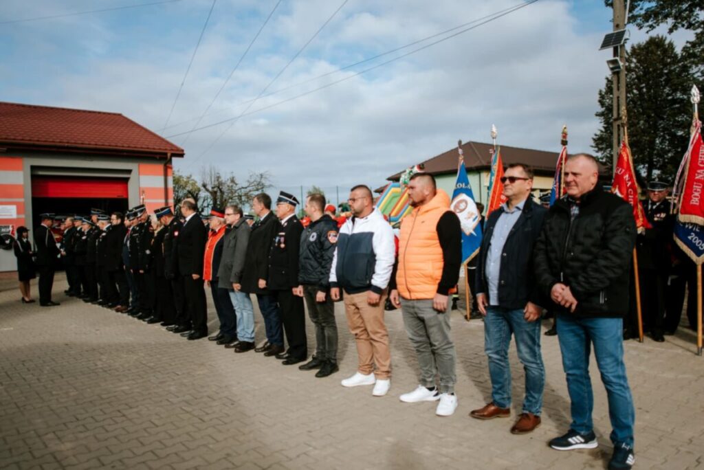 Grupa osób w strojach galowych i kurtkach stoi na placu brukowanym. Tło stanowi budynek z czerwonymi drzwiami i flagi. Niebo jest pochmurne.