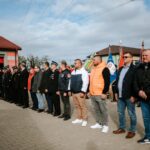 zUdWhBtP Grupa osób w strojach galowych i kurtkach stoi na placu brukowanym. Tło stanowi budynek z czerwonymi drzwiami i flagi. Niebo jest pochmurne.