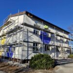 DInJURna Budynek z rusztowaniami, biały z niebieskimi i żółtymi elementami. Na rusztowaniu pracownik w zielonej kurtce. Tło to jasne, niebieskie niebo.