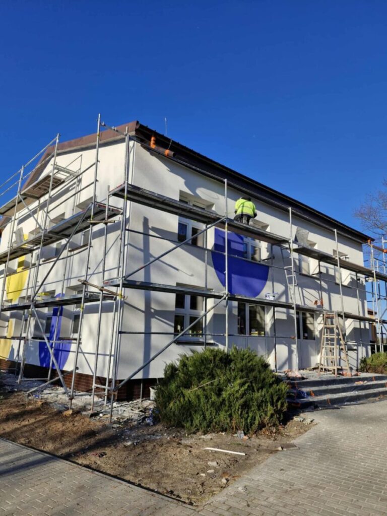 Budynek z rusztowaniami, biały z niebieskimi i żółtymi elementami. Na rusztowaniu pracownik w zielonej kurtce. Tło to jasne, niebieskie niebo.