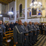 Wnętrze kościoła z żołnierzami w szarych mundurach na pierwszym planie. Tło ozdobione malowidłami i wiszącymi żyrandolami. W ławkach siedzą ludzie, drewniana podłoga z biało-czarnymi