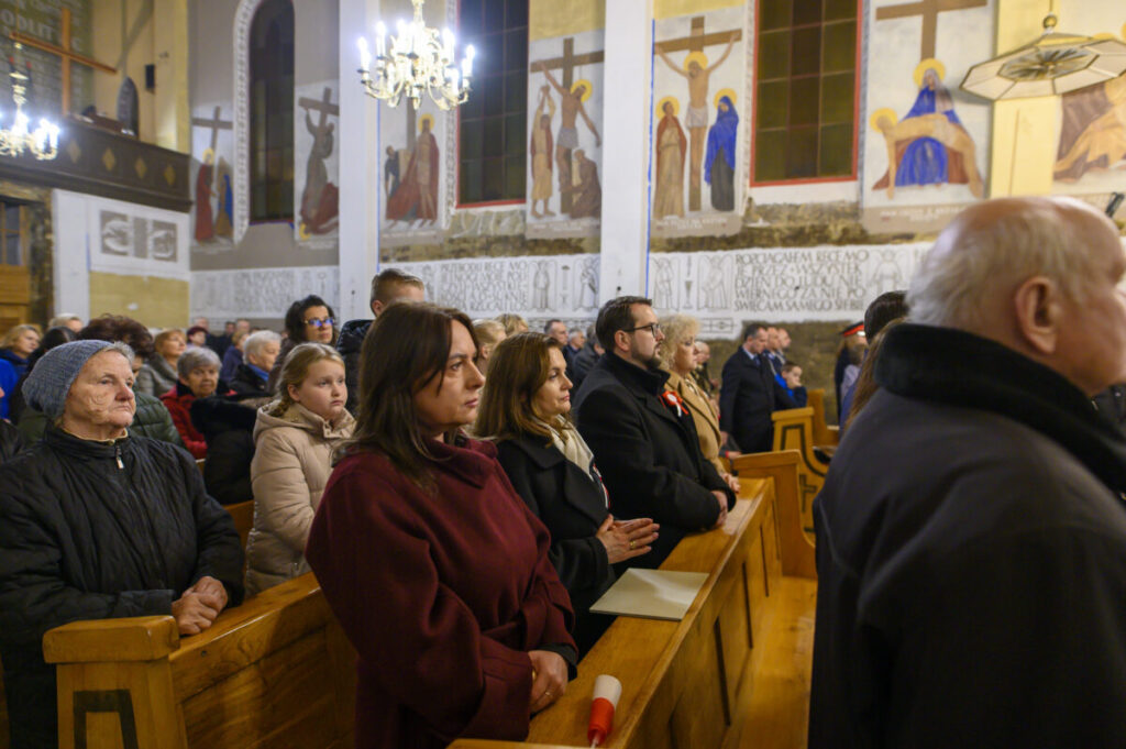 Grupa ludzi w kościele siedzi na drewnianych ławkach. Ściany zdobią kolorowe freski z biblijnymi scenami. Wnętrze oświetlają kryształowe żyrandole.