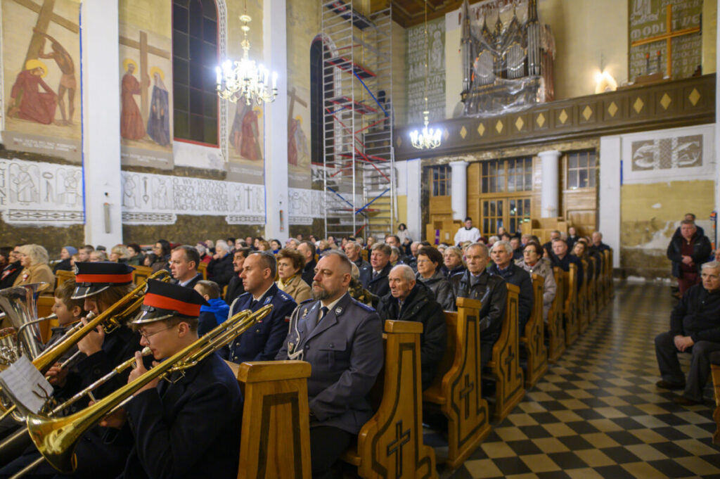 Wnętrze kościoła, pełne ludzi siedzących na ławkach. Ściany ozdobione malowidłami, sufit zdobi żyrandol. Na pierwszym planie muzycy z instrumentami.