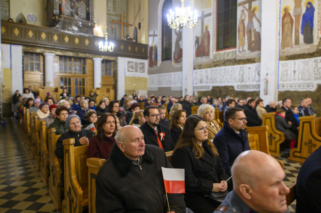 Obraz przedstawia ludzi siedzących w drewnianych ławkach wewnątrz kościoła. Wnętrze jest jasne, z ozdobnymi freskami na ścianach. Kobieta w ciemnym płaszczu trzyma polską flagę.